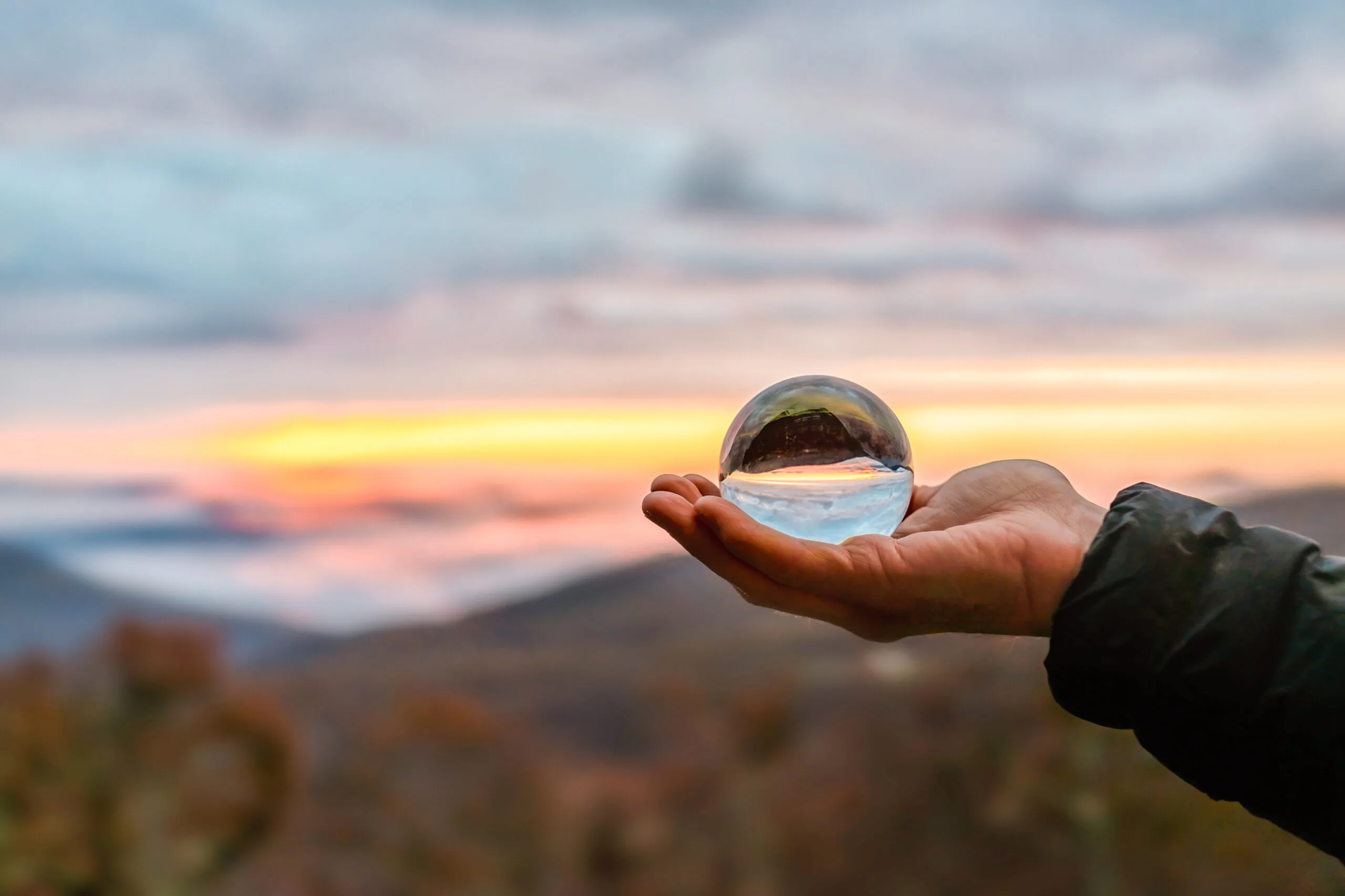 An open hand holding a glass sphere against a sunset over the hills