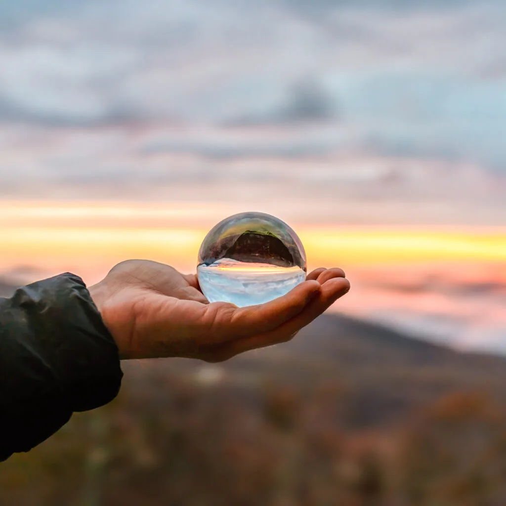 An open hand holding a glass sphere against a sunset over the hills
